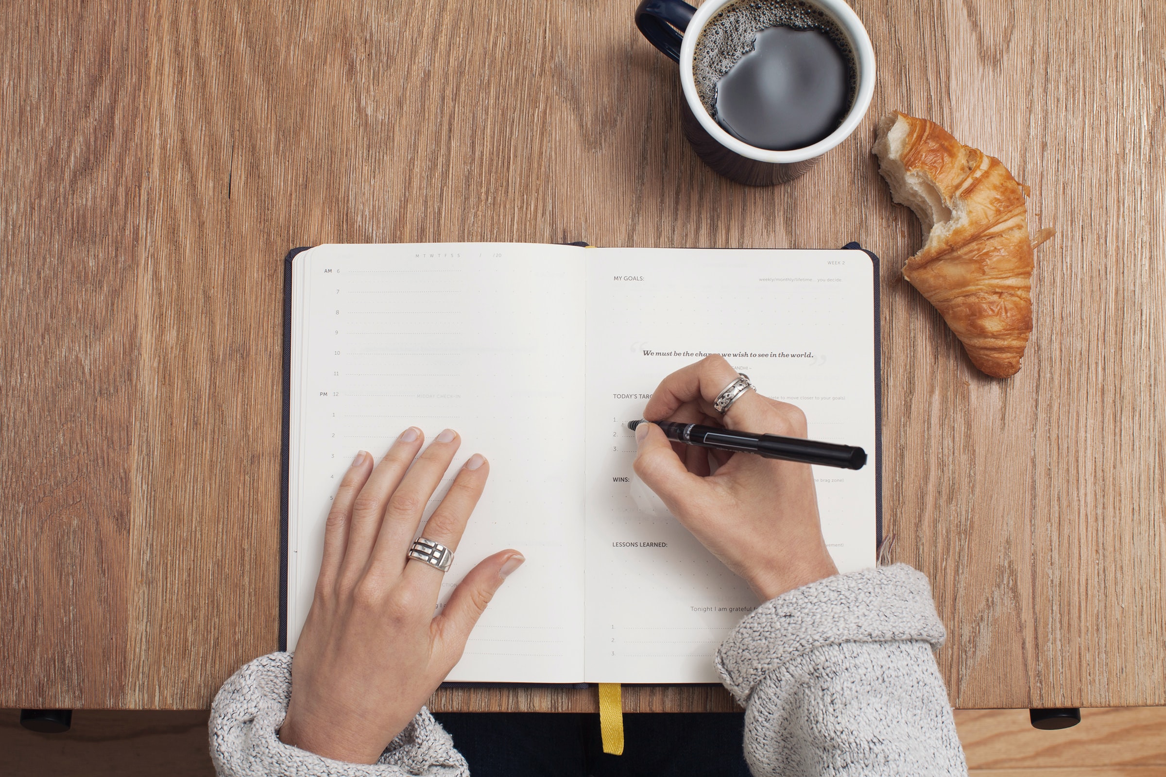 Woman achieving her goals and writing in her planner with coffee and croissant.